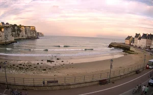 Vue réaliste du front de mer de Veules-les-Roses avec des vagues douces, une plage de sable, une promenade pittoresque et un ciel pastel au coucher du soleil.