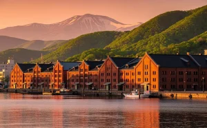 Les entrepôts en briques rouges de Hakodate longent un port calme devant des collines verdoyantes et des sommets enneigés, sous la lumière dorée du soir.